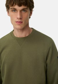 Jonge man met kort krullend bruin haar, gekleed in een olijfgroene crewneck trui, kijkt recht in de camera tegen een effen achtergrond.