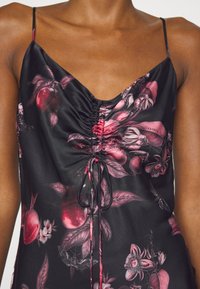 Femme portant une robe noire sans manches avec fines bretelles et motif floral et fruité rose, avec devant froncé et cordon noué.