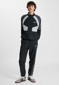 Ung man står med händerna i fickorna iförd en svart och grå hummel sportjacka och byxor, samt grå och vita sneakers.