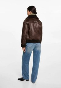 Jachetă din piele maro cu guler de blănă, cu manșete și tiv țesute, asociată cu blugi evazați de culoare albastră deschisă și bocanci negri până la gleznă.