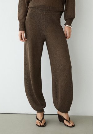 Losse bruine gebreide broek met omgeslagen enkels, gedragen met bijpassende top en donkere sleehakken op een lichte vloer- en muurachtergrond.