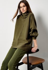 Pull en maille oversized vert olive avec un col montant et des manches ballons. Associé à un pantalon ajusté vert assorti, assis sur un tabouret en bois.