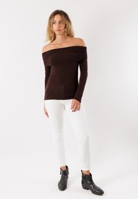 Top marrone off-the-shoulder con dettagli in tessuto arricciato, maniche lunghe, abbinato a pantaloni bianchi a vestibilità slim e stivaletti neri.
