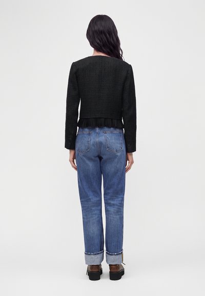 Γυναίκα που στέκεται πλάτη, φοράει μαύρο cropped jacket με μακρύ μανίκι, μπλε τζιν με γυρισμένα μπατζάκια και καφέ πλατφόρμες σε απλό φόντο.