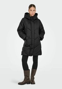 Cappotto piumino nero con cappuccio, dotato di un motivo a chevron trapuntato, tasche laterali e una lunghezza fino al ginocchio, indossato sopra leggings neri e stivaletti marroni.