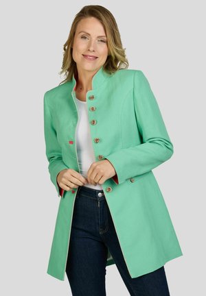 Vrouw draagt een muntgroene jasje met knopen over een wit shirt en donkere spijkerbroek, glimlacht zacht tegen een effen lichte achtergrond.