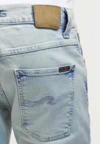 Light blue denim jeans back pocket with wave stitching and brown leather brand patch above the waistband.