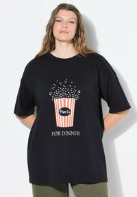 Camiseta negra de gran tamaño que presenta un gráfico de un cubo de palomitas con rayas rojas y blancas, texto que dice "Perlas PARA CENAR" y detalles con puntos.