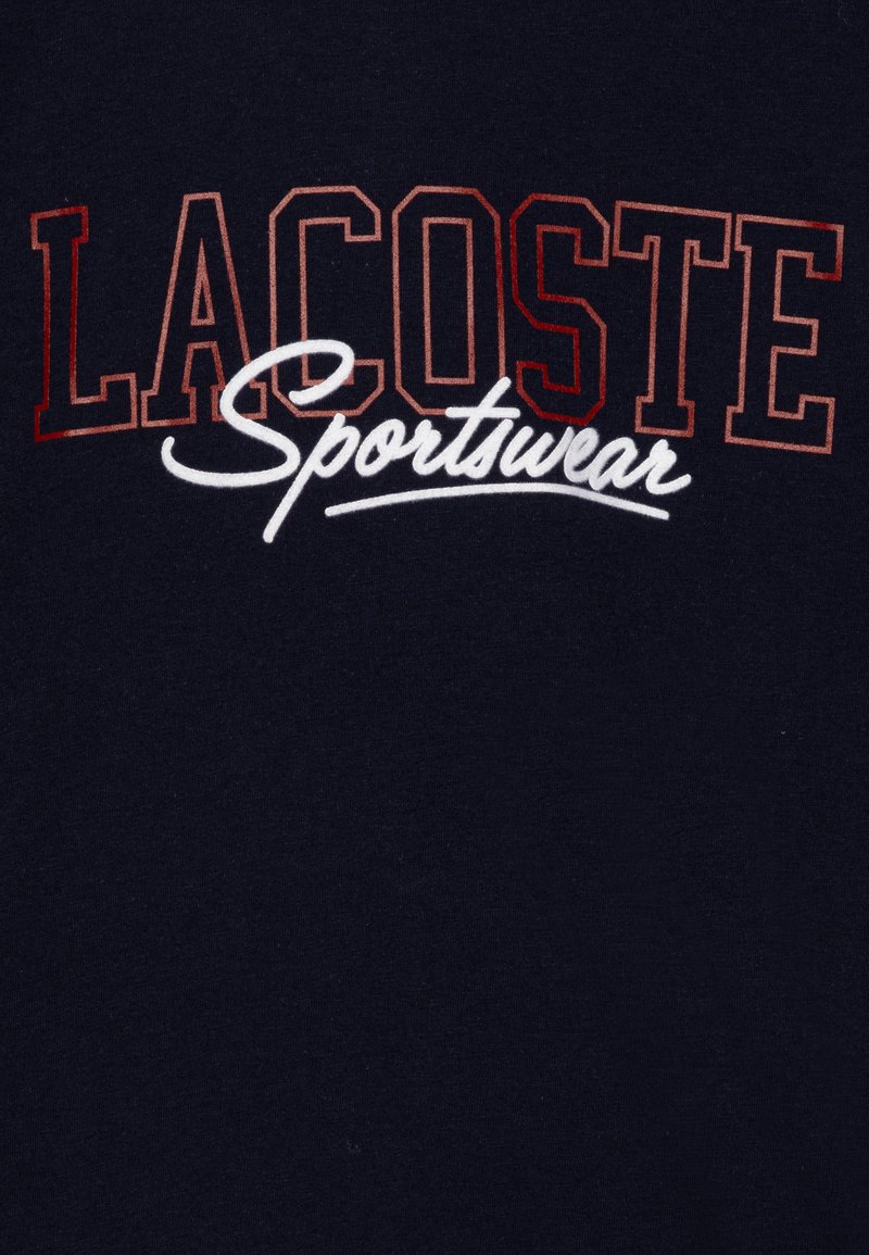 T-shirt navy con "LACOSTE" in lettere rosse in grassetto e "Sportswear" in corsivo bianco. Tessuto morbido, design casual, adatto per l'uso quotidiano.