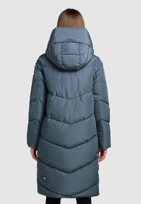 Langer Steppmantel in gedämpftem Türkis, mit hohem Kragen, Chevron-Steppmuster und einem Logodetail auf der unteren rechten Seite.