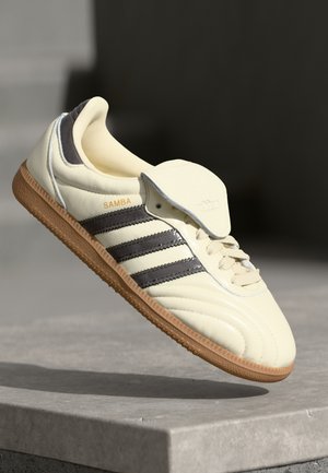 Sneaker de couro creme com três riscas escuras, sola de borracha castanha e bico arredondado. Apresenta uma língua dobrável rotulada como "SAMBA."