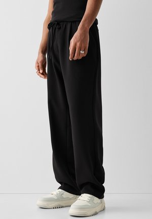 Mężczyzna w luźnych czarnych spodniach z wiązaniem w pasie, białych trampkach i srebrnych pierścieniach na palcach, stojący na tle jasnej, gładkiej ściany.