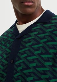 Muž oblečený v kardiganu s geometrickým vzorem v zeleno-námořnických barvách, s knoflíky, přes bílé tričko s kulatým výstřihem, zkrácené na krku a ramenou.