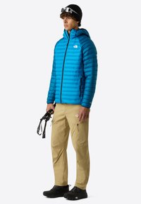 Blå puffjacka med horisontell quiltning, fickor med blixtlås och huva, ihop med beige cargobyxor och svarta skor. Individen håller klättringsutrustning.