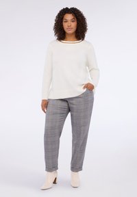 Suéter blanco con detalles de cadena en el escote. Pantalones de cuadros grises, ajustados. La modelo lleva botas de tobillo beige. Textura de tela suave.