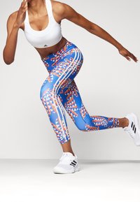 Urheilulegginsit sinisessä, joissa on geometrisia kuvioita punaisena ja valkoisena. Sisältävät kolme valkoista raitaa sivuilla. Materiaali vaikuttaa joustavalta.