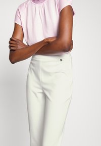 Jasnoróżowy sweter z krótkim rękawem i marszczonym dekoltem, zestawiony z białymi, dopasowanymi spodniami z bocznym rozcięciem i ozdobnym guzikiem.