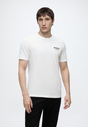 Ung mand med kort mørkt hår iført en hvid Hugo-branded t-shirt og sorte bukser, der står mod en ensartet lys baggrund.