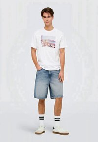 T-shirt branca com uma estampa gráfica, combinada com calções de ganga azul claro, meias brancas com riscas pretas e sapatilhas brancas.