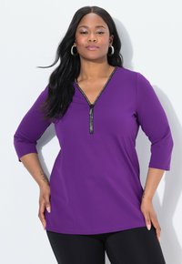 Paarse top met een V-hals en driekwartmouwen, voorzien van een ritssluiting aan de voorkant en metallic accenten. Zachte stof met een gladde textuur.