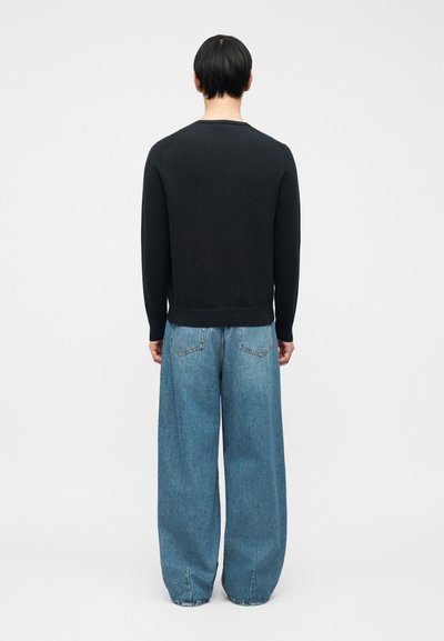 Άτομο με πλάτη, φορώντας μακρυμάνικο μαύρο πουκάμισο και άνετο μπλε τζιν, μπροστά σε απλό λευκό φόντο.