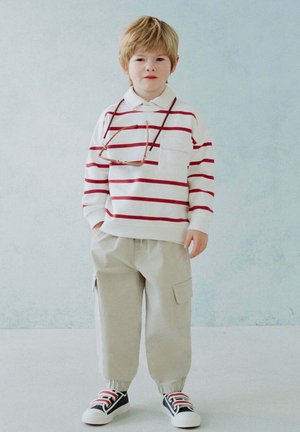 Jeune enfant aux cheveux blonds portant une chemise blanche à rayures rouges, un pantalon cargo beige, des baskets et des lunettes suspendues autour du cou.