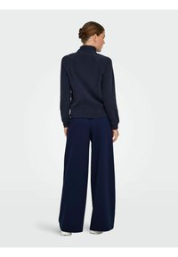 Tamno plava flis majica s visokim ovratnikom; uparen s širokim tamnoplavim hlačama. Glatka tekstura, duži rukavi i opušteni kroj.