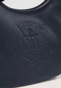 Marinblå läderväska med präglad logodetalj med ankare och banner. Slät textur, rund form och dragkedjestängning.