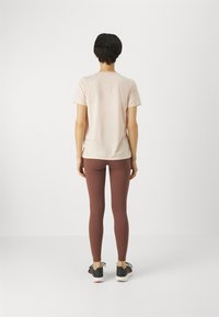 Person stående med ryggen mot kameran, iförd en ljusrosa kortärmad skjorta, bruna leggings och svarta sneakers med vita sulor.