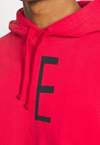 Sudadera roja con una impresión de la letra "E" en negro en la parte frontal. Cuenta con capucha ajustable y puños de canalé. Tejido de algodón suave.