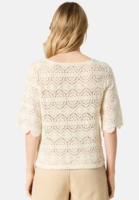 Blusa di pizzo color crema con maniche corte a campana e orlo scallop. Presenta motivi geometrici e un design a punto aperto.