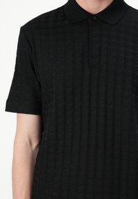 Svart kortermet polo skjorte med et teksturert mønster, tradisjonell krage og knappestolpe. Stoffet ser mykt og strukturert ut.