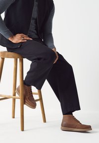 Bottes marron à lacets avec une tige en cuir lisse, des coutures contrastantes et une semelle texturée, portées avec un pantalon noir et un gilet zippé.