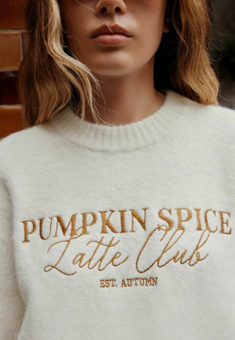 Woman wearing a white sweater with orange text reading "PUMPKIN SPICE Latte Club EST. AUTUMN" visible from shoulders to lips.