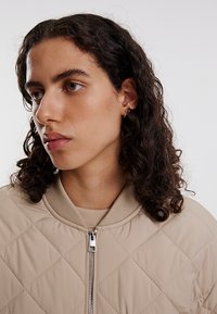 Beige gesteppte Bomberjacke mit einem Reißverschluss und geripptem Kragen. Das Design zeichnet sich durch Rautensteppung und eine glatte Textur aus.