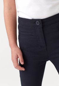 Pantaloni blu navy in tessuto morbido, con tasche frontali, chiusura a bottone e vestibilità sartoriale. Ingrandimento della vita e posizione della mano.