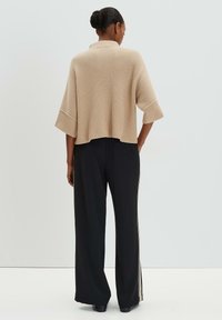 Beige gerippter Strickpullover mit übergroßen Ärmeln, kombiniert mit schwarzen weit geschnittenen Hosen mit seitlichen Streifen. Ansicht von hinten.