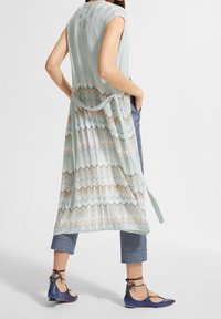 Gilet lungo senza maniche di maglia azzurro chiaro con motivo a zigzag, legato in vita, indossato sopra pantaloni grigi e abbinato a scarpe basse blu scuro.