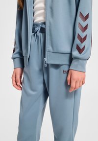 Jasnoniebieska kurtka na zamek i dopasowane spodnie wykonane z gładkiego materiału. Kurtka ma bordowe akcenty w zygzak na rękawach oraz logo na spodniach.