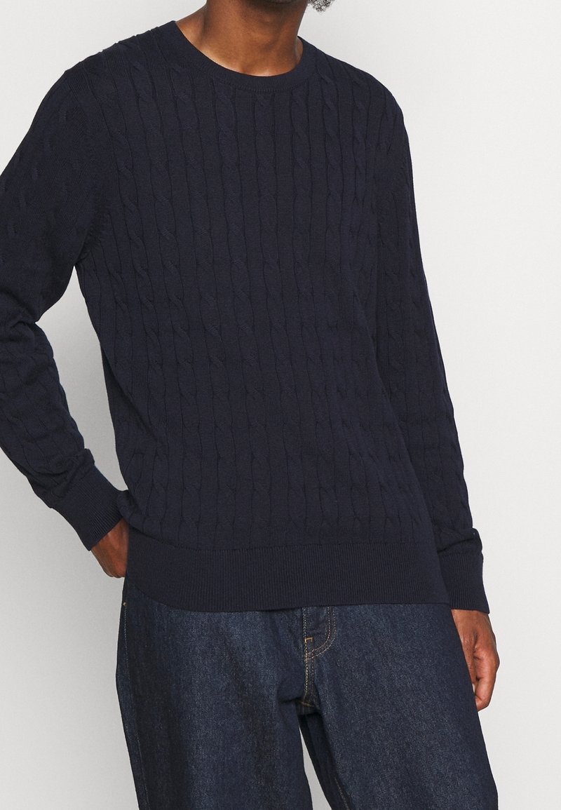 Homme portant un pull en maille torsadée bleu marine et un jean bleu foncé, debout avec une main dans la poche devant un fond uni.