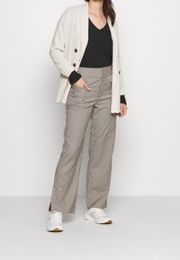 Beżowa marynarka z brązowymi guzikami, czarna bluzka z dekoltem w serek i w kratkę spodnie. Białe sneakersy z jasnoszarymi i beżowymi akcentami. Jednolity tło.