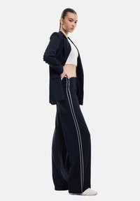 Marinblå kavaj över en vit crop top, kombinerad med vida marinblå byxor med vita sidostråk. Slätt tyg med strukturerad design.