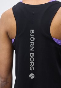 Κοντινή εικόνα από το άνω μέρος της πλάτης ενός ατόμου που φοράει μαύρο αμάνικο τοπ Björn Borg πάνω από μοβ αθλητικό σουτιέν.