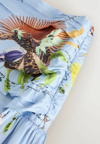 Tissu bleu clair avec un imprimé vibrant d'oiseaux tropicaux et de feuillage, avec une écriture de style manuscrit et des détails froncés et plissés.