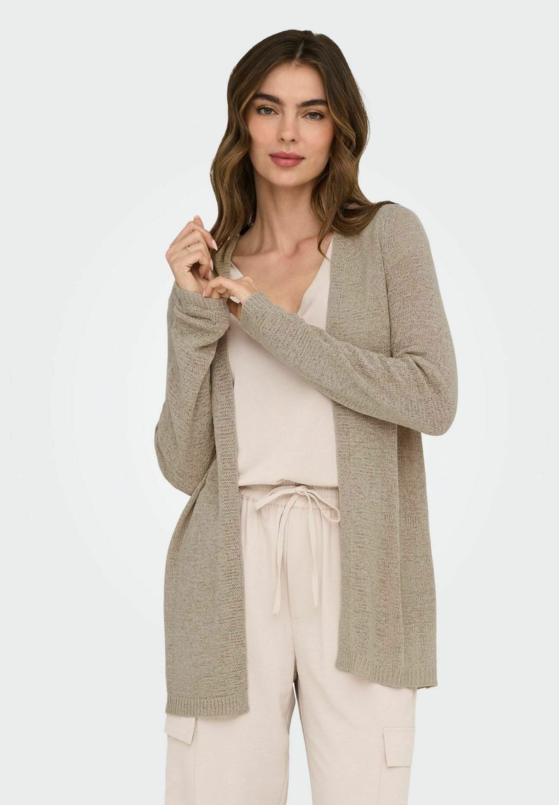 Gilet en tricot beige avec un devant ouvert, des manches longues et des poignets côtelés. Assorti à un haut de couleur claire et à un pantalon, créant un look superposé.