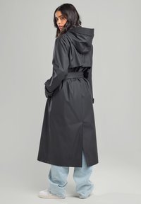 Long black waterproof coat with a hood, belted waist, and back slit. Worn over light blue baggy jeans and white sneakers. Smooth texture.