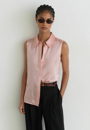 Vrouw met gevlochten haar, die zwarte zonnebrillen, een lichtroze mouwloze blouse en zwarte broek met hoge taille draagt, staand tegen een witte muur.