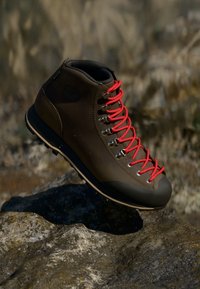 Brun turstøvel med høy topp-design, røde snører, metalløye og en teksturert gummisåle, plassert på en steinoverflate.