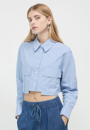 Chemise bleu rayée coupée en tissu léger, avec un devant à boutons, deux poches poitrine et de longues manches. Associée à un short en denim.