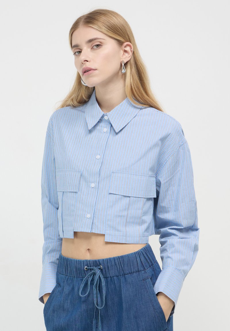 Gekürzte blau gestreifte Bluse aus leichtem Stoff, mit Knopfleiste, zwei Brusttaschen und langen Ärmeln. Kombiniert mit Jeansshorts.